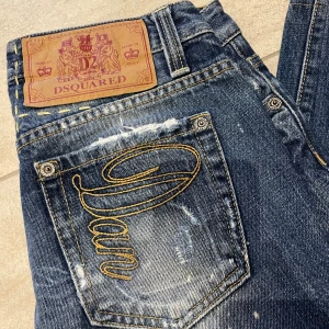 Dsquared2 blå jeans med broderad ficka - Säljer ett par blå jeans från Dsquared2 med coolt broderi på bakfickorna och slitna detaljer. Jeansen har klassisk femficksdesign, raka ben och en snygg läderpatch bak i midjan. Materialet är robust denim med gul kontrastsöm. Storlek 28/30