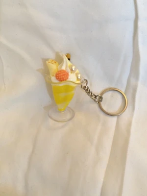 Nyckelring sundae - Bilderna visar en nyckelring formad som en gul och vit glassdessert med dekorativa pärlor och en liten orange detalj.