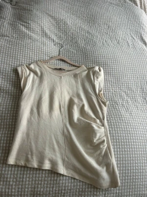 Beige ärmlös topp från Zara - Säljer en stilren beige topp från Zara i storlek M. Toppen är ärmlös med rund hals och har en rak passform. Materialet är mjukt och känns som bomull, perfekt för varma dagar. Snygg söm framtill som ger en cool detalj.