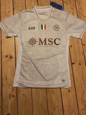 Napoli tröja - Bilderna visar en vit fotbollströja från Napoli med subtila mönster, bruna loggor och detaljer, samt texten 'Proud to be Napoli' på ryggen. 