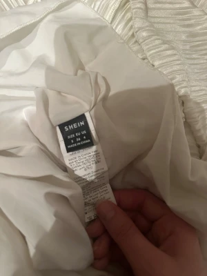 Vit plisserad långklänning från SHEIN - Säljer en elegant vit långklänning från SHEIN i storlek S. Klänningen har plisserat tyg med skimrande finish och långa ärmar. Materialet är syntet och ger en lyxig känsla. Perfekt för dig som gillar stilrena och trendiga looks.