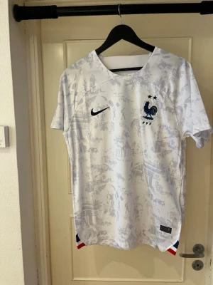 France away shirt 22 - Säljer en officiell Nike Frankrike fotbollströja i storlek L. Tröjan är vit med ljusblå mönster av franska landmärken och har broderad tupp-logga med två stjärnor på bröstet. Dri-FIT material för extra komfort och svetttransport. Om du vill ha eget tryck och namn där bak hör av dig! MVH Moncliqué 🥂