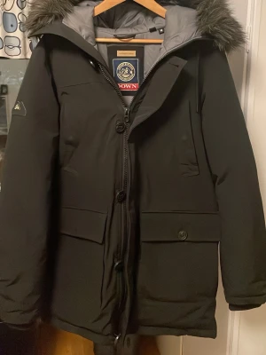 Svart dunjacka Superdry med päls - Säljer en svart dunjacka från Superdry med huva och päls. Jackan har flera fickor, dragkedja och knappar framtill samt grått innerfoder. Perfekt för kalla dagar och riktigt snygg streetstil. Köpt för 3000kr. Priset är diskuterbart!