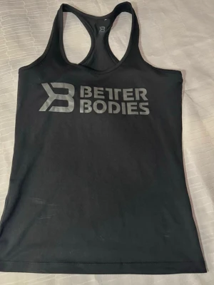 Svart Better Bodies linne M - Säljer en svart linne  från Better Bodies i storlek M. Toppen har ett sportigt racerback-rygg och ett stort tryck med Better Bodies-loggan framtill. Materialet är mjukt och stretchigt, perfekt för gymmet eller annan träning.