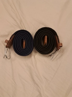 Två flätade bälten i svart och marinblå - Säljer två stilrena flätade bälten, ett i svart och ett i marinblått. Båda har detaljer i brunt konstläder med vit söm och silvrigt spänne. Perfekta för att lyfta vilken outfit som helst med en klassisk touch. Har aldrig använt! Kna diskussion pris, tveka inte att skriva om du har några frågor!