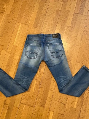 Replay blå jeans - Säljer ett par Replay Blue Jeans i klassisk blå tvätt med snygga slitningar och raka ben. Jeansen har fem fickor, normal midja och tydliga Replay-detaljer på bakfickorna. Perfekta till vardags för dig som gillar en avslappnad men stilren look. 