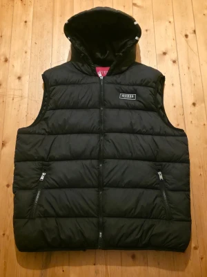 Svart dunväst med huva från Guess - Säljer en svart dunväst från Guess med stor huva och dragkedja framtill. Västen har två fickor med dragkedja och Guess-logga både på bröstet och på huvan. Nypris runt 3000kr. Passar XL