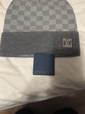 Grå rutig Louis Vuitton mössa - Snygg grå mössa från Louis Vuitton med rutigt mönster upptill och enfärgad uppvikt kant. LV-logga broderad på kanten. Tillverkad i mjuk ullblandning som känns lyxig och varm. Perfekt accessoar för att lyfta din vinterstil.