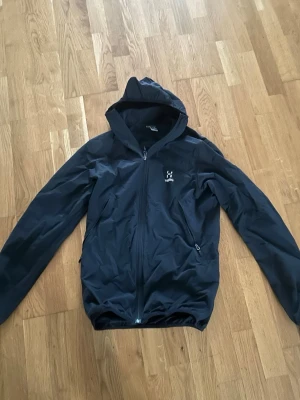 Svart vindjacka från Haglöfs - Säljer en svart vindjacka från Haglöfs med huva och hel dragkedja framtill. Jackan har två sidofickor med dragkedja och en enkel, sportig design. Perfekt för blåsiga dagar och outdoor-äventyr.