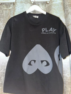 Svart Comme des Garçons PLAY t-shirt - Svart t-shirt från Comme des Garçons PLAY med stora gråa hjärtögon-prints fram och bak. Klassisk rund hals och korta ärmar. Tryckt PLAY-logga på bröstet. Tillverkad i mjuk bomull för skön känsla och stilren look. Oversize