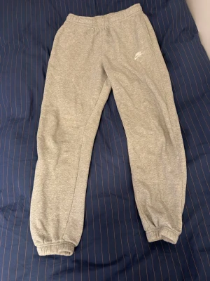 Grå mjukisbyxor från Nike - Säljer ett par klassiska grå mjukisbyxor från Nike med resår i midjan och vid bensluten. Byxorna har en enkel och clean look med Nike-logga tryckt på vänster sida. Perfekt för chill dagar eller när du vill vara bekväm.
