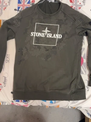 Grön sweatshirt från Stone Island - Säljer en grön sweatshirt från Stone Island med stort vitt tryck och logga på bröstet. Tröjan har ett diskret rutmönster i olika nyanser och ribbade muddar vid ärmslut och nederkant. Perfekt för dig som gillar streetwear och vill ha något stilrent.