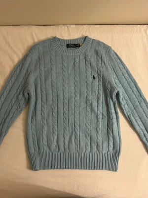 Ljusblå kabelstickad tröja Polo Ralph Lauren - Säljer en ljusblå kabelstickad tröja från Polo Ralph Lauren. Tröjan har rund hals, ribbade muddar och det klassiska broderade logot på bröstet. Perfekt för dig som gillar preppy stil och vill ha något både snyggt och bekvämt. (Oanvänd)