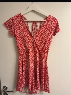 Röd blommig omlott playsuit - Superfin röd playsuit med vita blommor och omlottmodell. Klänningen har korta ärmar, v-ringning och knytning i ryggen. Perfekt för varma dagar och ger en söt och somrig vibe.