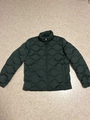 Grön quiltad jacka från Dressmann – strl M - Stilren och varm quiltad jacka från Dressmann. Mörkgrön färg som passar till det mesta. Perfekt för höst, vinter och tidig vår. Jackan har dragkedja framtill, två sidofickor med dragkedja och en bekväm krage.                                     Använd men i skick som ny, inga hål eller fläckar. Säljs eftersom den inte längre används.                Nypris 1400kr 📦 Skickas snabbt 🏠 Djurfritt och rökfritt hem 