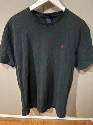 Svart t-shirt från Polo Ralph Lauren - Klassisk svart t-shirt från Polo Ralph Lauren med röd broderad logga på bröstet. Tillverkad i 100% bomull och har en normal passform med korta ärmar och rund hals. Perfekt basplagg för en clean och enkel stil. Nypris 999 pris 149