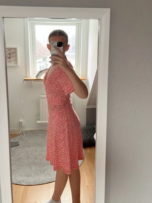 Röd blommig kortklänning med v-ringning - Superfin röd kortklänning med vitt blommönster och v-ringad hals. Klänningen har korta ärmar och en lätt utställd kjol som ger en skön och luftig känsla. Perfekt för varma dagar och enkel att styla med sneakers eller sandaler.