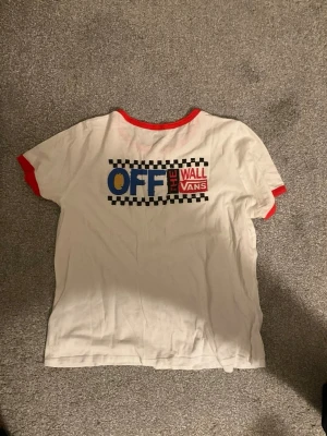 Vit Vans t-shirt med röd kant - Vit t-shirt från Vans med röda kanter vid hals och ärmslut. Tryck med Vans-logga fram och stort 'Off The Wall' tryck bak med schackrutigt mönster i svart, blått och rött. Klassisk och streetinspirerad stil, perfekt för dig som gillar skatekulturen. Köptes i USA för 400 kr.