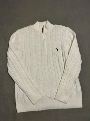 Vit kabelstickad tröja Polo Ralph Lauren - Vit kabelstickad tröja från Polo Ralph Lauren med halv dragkedja och klassisk broderad logga på bröstet. Tröjan har ribbade muddar, hög krage och är långärmad. Perfekt för dig som gillar stilrena och tidlösa plagg.