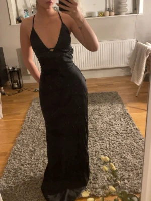 Svart balgklänning med öppen rygg - Säljer en svart långklänning med djup v-ringning och tunna axelband som korsas och knyts i ryggen. Klänningen har en figurnära passform och en elegant, minimalistisk look. Perfekt för dig som vill ha en stilren och snygg outfit.