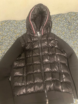 Svart Moncler puffar cardigan med huva - Säljer en svart pufferjacka från Moncler med glansig yta och ribbstickade ärmar. Jackan har huva med röd och vit detalj, dragkedja framtill och två fickor. Perfekt för kyliga dagar och riktigt snygg streetstil.  Jag säljer denne pga att den är större i midjan och jag har köpt en ny till mig. Märket går att scanna. Pris kan även diskuteras 