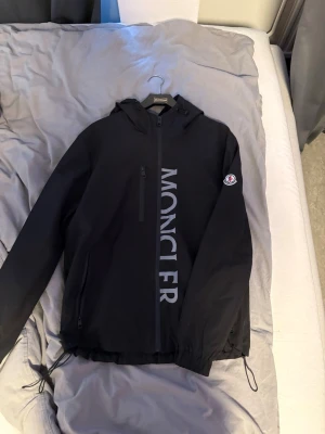 Svart vindjacka från Moncler - Säljer en svart vindjacka från Moncler med stor logga framtill och klassisk Moncler-patch på ärmen. Jackan har huva, dragkedja, snörning i nederkant och flera fickor. Perfekt för dig som vill ha en stilren och sportig look.