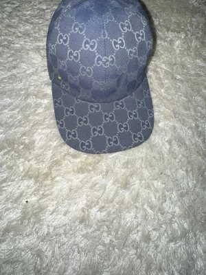 Blå Gucci keps  - Säljer en blå keps från Gucci med klassiskt GG-monogram över hela kepsen. Justerbar rem bak i matchande blått och insida i beige bomull. Snygg och stilren accessoar som lyfter vilken outfit som helst.