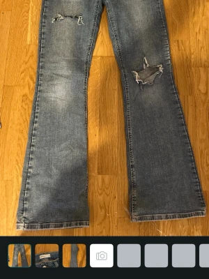 Blå bootcut jeans med slitningar - Säljer ett par blå jeans med bootcut passform och tydliga slitningar på båda benen. Jeansen har en klassisk femficksdesign och är tillverkade i jeansmaterial. Perfekta för en avslappnad och trendig look.
