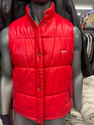Röd dunväst från Story - Snygg röd dunväst från Story med quiltad design och hög krage. Västen har tryckknappar framtill, dragkedjefickor och broderad logga på bröstet. Fodrad med Dacron Hollofil för extra värme. Perfekt för lager på lager under kyliga dagar.