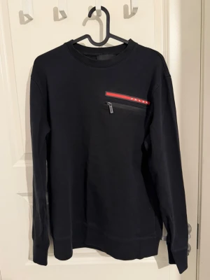 Svart sweatshirt från Prada - Säljer en svart sweatshirt från Prada med röd logga och dragkedjeficka på bröstet. Tröjan har rund halsringning och långa ärmar. Perfekt för dig som vill ha en clean och exklusiv look med streetvibe.