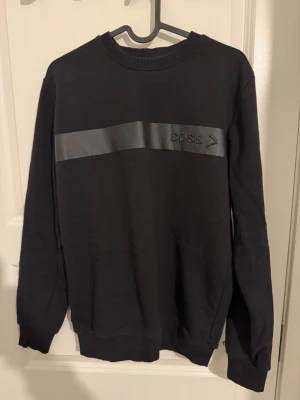 Svart boss tröja - Säljer en svart sweatshirt från BOSS i storlek S. Tröjan har en bred, blank rand över bröstet med präglad BOSS-logga. Klassisk rund halsringning och långa ärmar. Perfekt för dig som gillar stilrena och snygga plagg.