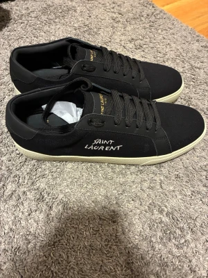 Svarta Saint Laurent skor - Säljer dessa svarta Saint Laurent skor med vit broderad logga på sidan och gulddetaljer på plösen. Skorna har snörning, vit platt sula och är tillverkade i textil och skinn. Klassisk och stilren modell som passar till typ allt. 