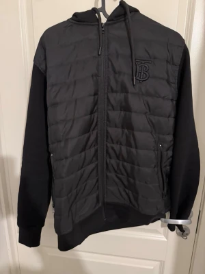 Svart quiltad pufferjacka Burberry - Säljer en svart quiltad pufferjacka från Burberry med dragkedja framtill, huva med snörning och broderad TB-logga på bröstet. 