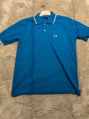 Blå pikétröja från Fred Perry - Säljer en klassisk blå pikétröja från Fred Perry med vit logga broderad på bröstet. Tröjan har vit rand på krage och ärmslut, samt knappar framtill. Perfekt till en avslappnad men ändå stilren look.