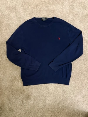 Ralph Lauren Marinblå Stickad Tröja - Storlek L - Slim Fit - 100% Bomull - Säljen en marinblå stickad tröja från Polo Ralph Lauren i lyxig 100% Pima bomull. Klassisk crew neck-design med det ikoniska röda polo-märket broderat på bröstet. Slim Fit-modell som ger en modern, snygg silhuett. Perfekt till både vardags och finare tillfällen! Detaljer:  Märke: Polo by Ralph Lauren Storlek: L (Large) - Slim Fit Färg: Marinblå/Navy Blue Modell: Stickad tröja/Sweater med rund hals Skick: Mycket gott - använd men välvårdad Material: 100% Pima Cotton (exklusiv premium bomull) 
