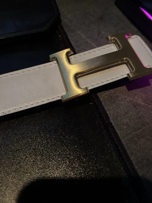 Vit skärp med guldspänne H - Säljer ett stilrent vitt skärp med ett stort, blankt H-format spänne i guld. Skärpet har slät yta och tydliga sömmar längs kanterna. Perfekt accessoar för att lyfta din outfit med en lyxig touch.