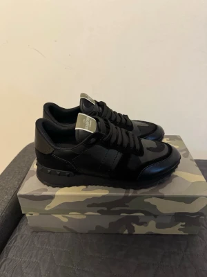 Valentino Garavani Rockrunner sneakers - Säljer ett par svarta Valentino Garavani Rockrunner sneakers med coola kamouflagemönster och mattsvarta detaljer. Skorna har snörning, robust sula med nitar och är tillverkade i en mix av skinn och textil. Perfekta för dig som vill ha en lyxig och edgy sneaker-look.