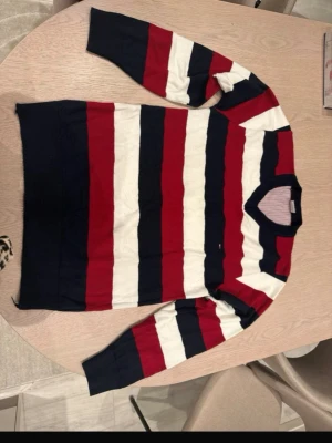 Randig tröja Tommy Hilfiger M - Säljer en klassisk stickad tröja från Tommy Hilfiger i storlek M. Tröjan har breda marinblå, röda och vita ränder, rund halsringning med ribbad kant och diskret logga på bröstet. Perfekt för dig som gillar preppy stil och vill ha något som sticker ut.