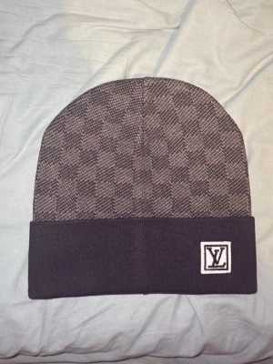 Rutig mössa från Louis Vuitton - Säljer en snygg och exklusiv mössa från Louis Vuitton i mörkgrått och svart med klassiskt rutigt mönster. Mössan har vikt kant med LV-logga framtill och är tillverkad i mjukt material som känns lyxigt och bekvämt.