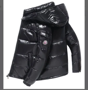 Svart pufferjacka från Moncler - Säljer en svart, glansig pufferjacka från Moncler med huva och dragkedja. Jackan har Moncler-logga på ärmen, flera fickor och quiltad design. Perfekt för kalla dagar och riktigt snygg till streetwear-looken.