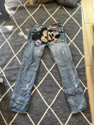 Blå jeans med stora patchar och brodyr - Unika blå jeans med slitningar, stora färgglada patchar och broderier på bakfickorna. Jeansen har ett rakt snitt och coola detaljer som applikationer i form av ansikte och text. Perfekta för dig som vill sticka ut med din stil.