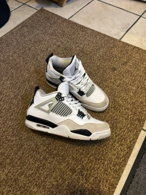 Nike Air Jordan 4  - Säljer ett par Nike Air Jordan 4 sneakers i vitt, svart och grått. Skorna har meshpaneler, snörning och detaljer i mocka och skinn. Klassisk siluett med synlig luftbubbla i sulan och ikoniska plastdetaljer på sidorna. Perfekta för dig som gillar streetwear och snygga sneakers.