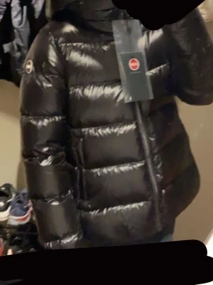 Svart pufferjacka från Colmar - Säljer en svart, glansig pufferjacka från Colmar med huva och dragkedja framtill. Jackan har en klassisk pufferlook med breda kanaler och logga på ärmen. Perfekt för kalla dagar och riktigt snygg till streetwear-stil.