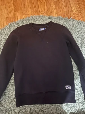 Svart sweatshirt från Jack & Jones - En stilren svart sweatshirt från Jack & Jones med rund hals och ribbade muddar. Tröjan har en enkel design med en liten logotyp nertill och är tillverkad i mjuk bomull. Perfekt för chill dagar eller när du vill ha en clean look.