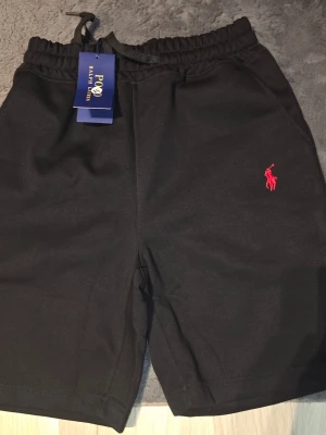 Svarta shorts från Polo Ralph Lauren - Svarta shorts från Polo Ralph Lauren med elastisk midja och snörning. Klassisk design med röd broderad logga på ena benet. Perfekta för en avslappnad stil och tillverkade i mjuk bomull för extra komfort.