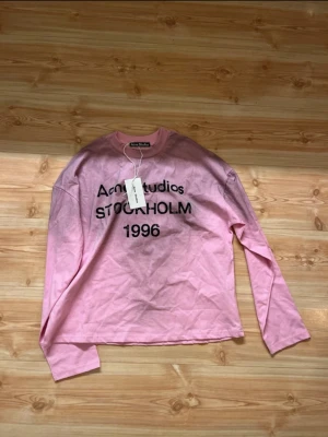 Rosa Acne Studios longsleeve - Säljer en rosa långärmad topp från Acne Studios med tryck i svart text 'Acne Studios STOCKHOLM 1996' på framsidan. Klassisk rund halsringning och avslappnad passform. Perfekt för dig som gillar streetwear och vill ha något stilrent men ändå statement.