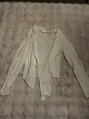 Vit omlottkofta från Lingon & Blåbär - Säljer en tunn, vit omlottkofta från Lingon & Blåbär i storlek M. Koftan har långa ärmar och är tillverkad i mjuk bomull. Perfekt att slänga över en topp för en enkel och clean look.