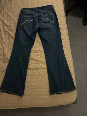 Blå bootcut jeans med kontrastsömmar - Säljer ett par blå bootcut jeans från Nelly med låg midja och snygga kontrastsömmar på bakfickorna. Jeansen har klassisk femficksdesign och är tillverkade i jeansmaterial med en mörk tvätt. Perfekta för dig som gillar retrovibbar och vill ha ett par riktigt schyssta jeans.