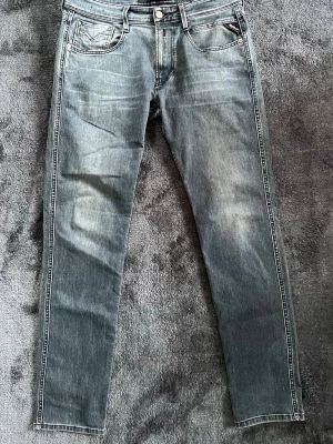 Replay Anbass jeans i blå denim - Säljer ett par Replay Anbass jeans i grå tvättad denim med klassiska fem fickor och snygga detaljer på bakfickorna. Jeansen har en rak passform och tydliga slitningar för en cool look. Perfekta för dig som gillar stilrena och bekväma jeans. Liten defekt i skrevet (bild 5)