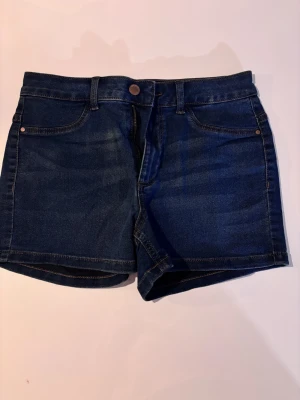 Mörkblå jeansshorts från JDY - Snygga mörkblå jeansshorts från JDY med klassisk femficksdesign, bälteshällor och dragkedja med knapp. Shortsen har en rak passform och är perfekta för varma dagar. Materialet är mjuk denim som känns bekvämt hela dagen.